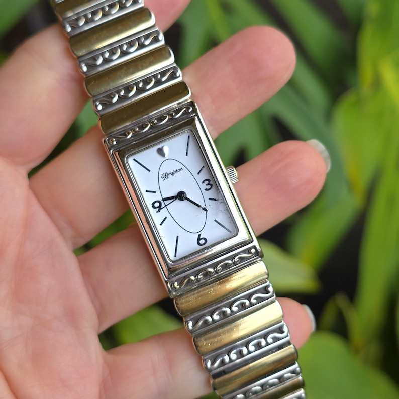 Op de afbeelding: Rechthoekig polshorloge met een witte wijzerplaat, zwarte wijzers en de cijfers 3, 6 en 9. Het horloge heeft een zilver- en goudkleurige band. De wijzerplaat heeft het woord "Brighton" en een klein hartsymbool.