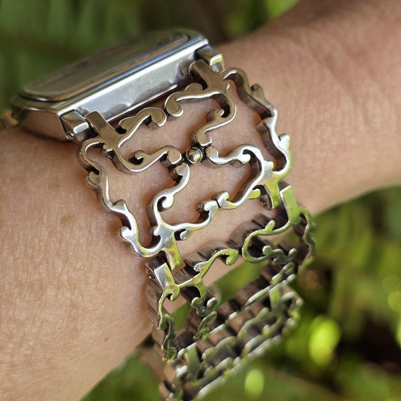 May include: A silver-toned watch with a rectangular face and an ornate, openwork bracelet. The bracelet features a repeating, scrolling design. The watch is worn on a wrist, with a blurred green background.