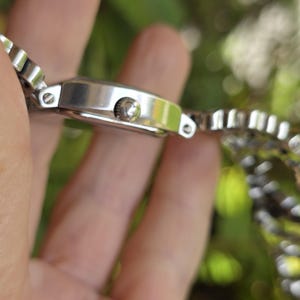 May include: A silver wristwatch with a rectangular face and a linked metal band. The watch has a small crown and a polished finish. The band is made of small, rectangular links.