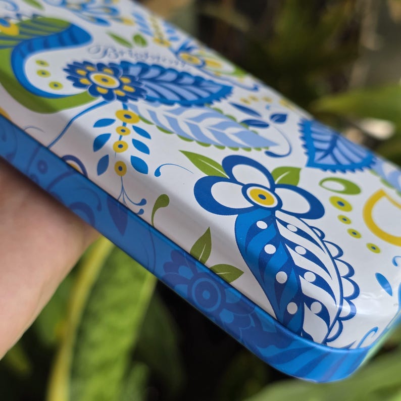 May include: A rectangular metal tin featuring a white background with a repeating pattern of blue and lime green floral and leaf motifs. The side of the tin is a solid blue with a subtle darker blue decorative pattern printed along the edge.