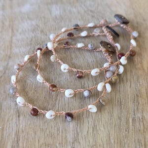 May include: Three handmade bracelets with a natural, bohemian aesthetic. Each bracelet features a series of small, white, brown, and gray beads strung on a light brown cord. The bracelets are fastened with wooden buttons.