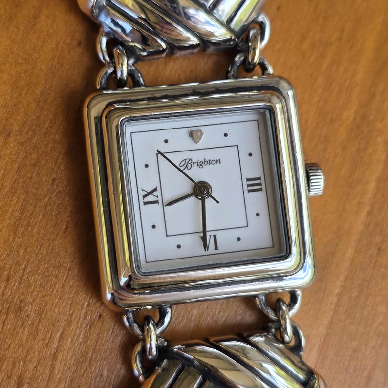 May include: A square-faced analog wristwatch with a white dial featuring the Brighton logo, Roman numerals at three, six, and nine o'clock, and a small heart icon at the twelve o'clock position. The watch has a polished silver-tone metal link band.