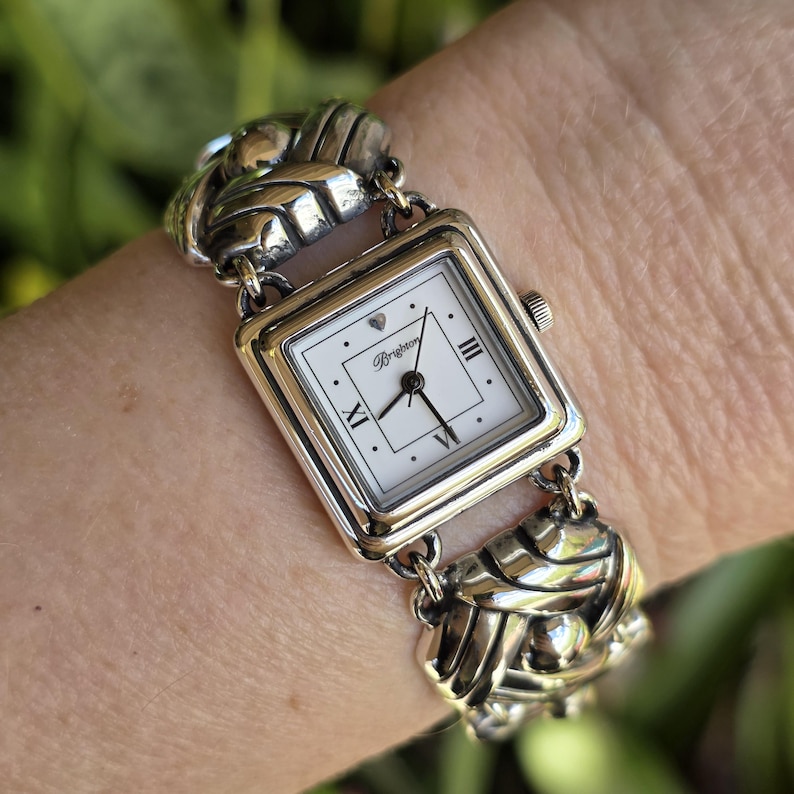 May include: A silver-tone analog wristwatch with a square white face featuring the Brighton logo, a small heart icon, and Roman numerals at the three, six, and nine o'clock positions. The band consists of interconnected, polished silver-tone metal links.