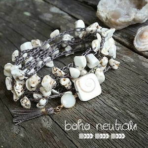 May include: A white and gray crocheted bracelet with white stone beads and a white shell charm. The bracelet has a gray tassel and is on a wooden surface. "boho neutrals" is written on the surface.