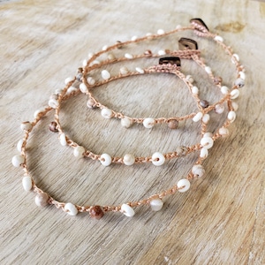 May include: Three handmade bracelets with a natural, bohemian aesthetic. Each bracelet features a woven, light brown cord adorned with small, white and tan beads. The bracelets are fastened with small, square wooden buttons.