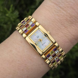 May include: A gold-toned wristwatch with a rectangular face and a band adorned with colorful gemstones. The watch face has a mother-of-pearl appearance with gold hands and Roman numeral hour markers. The watch is on a wrist.