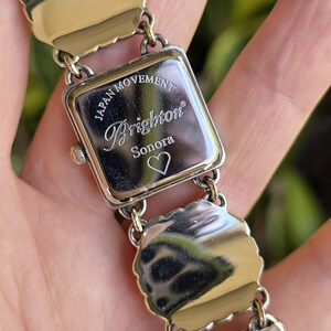 May include: A silver-tone metal wristwatch with a square case and a link bracelet. The back of the watch case is engraved with the text Japan Movement, Brighton, Sonora, and a heart symbol. The metal surfaces are polished and reflective.