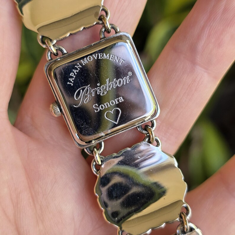 May include: A silver-tone metal wristwatch with a square case and a link bracelet. The back of the watch case is engraved with the text Japan Movement, Brighton, Sonora, and a heart symbol. The metal surfaces are polished and reflective.