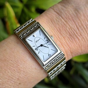 Op de afbeelding: Een rechthoekig zilverkleurig horloge met een witte wijzerplaat en zwarte cijfers. Het horloge heeft een zilveren band met decoratieve accenten. De wijzerplaat toont de merknaam "Brighton". Het horloge zit om een pols.