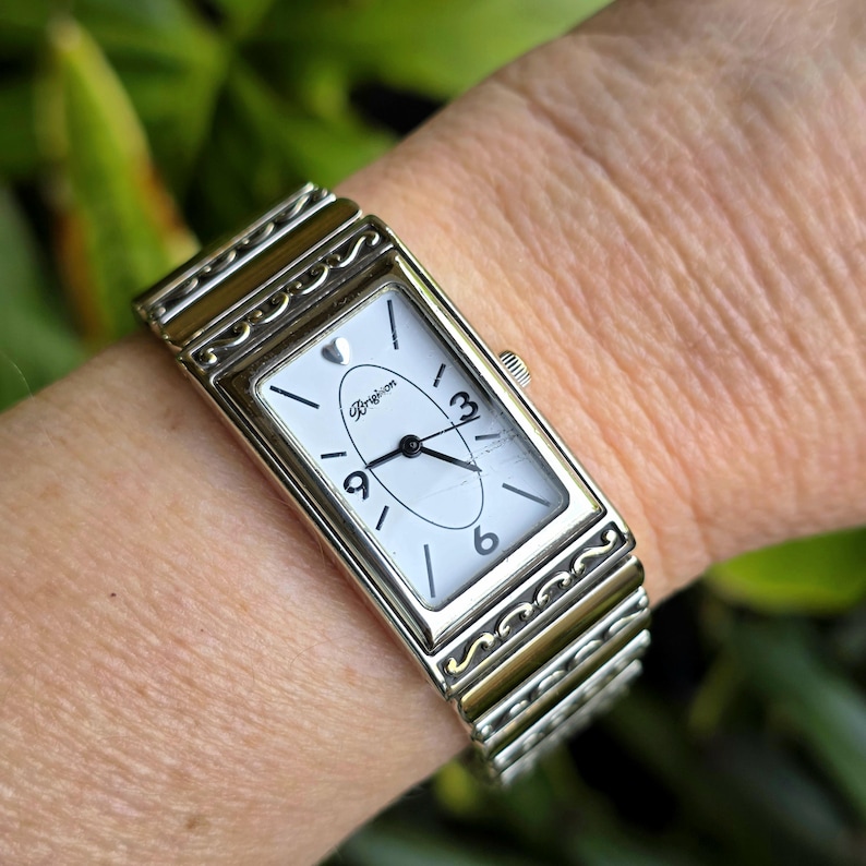 Op de afbeelding: Een rechthoekig zilverkleurig horloge met een witte wijzerplaat en zwarte cijfers. Het horloge heeft een zilveren band met decoratieve accenten. De wijzerplaat toont de merknaam "Brighton". Het horloge zit om een pols.