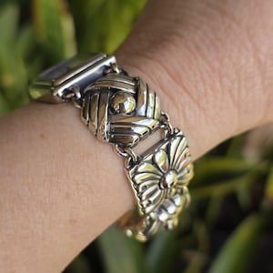 May include: A silver-toned watch featuring a link bracelet composed of square metal segments. Each segment displays a distinct embossed pattern, including a woven knot design and a floral motif, connected by small circular metal links.