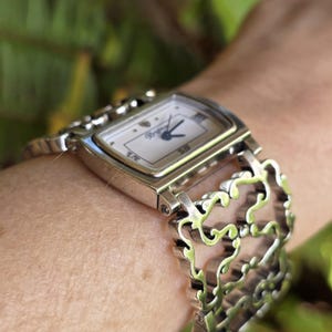 May include: A silver-toned wristwatch with a rectangular face and a decorative, open-link band. The watch face is white with black hands and the brand name "Brighton". The watch is worn on a wrist.