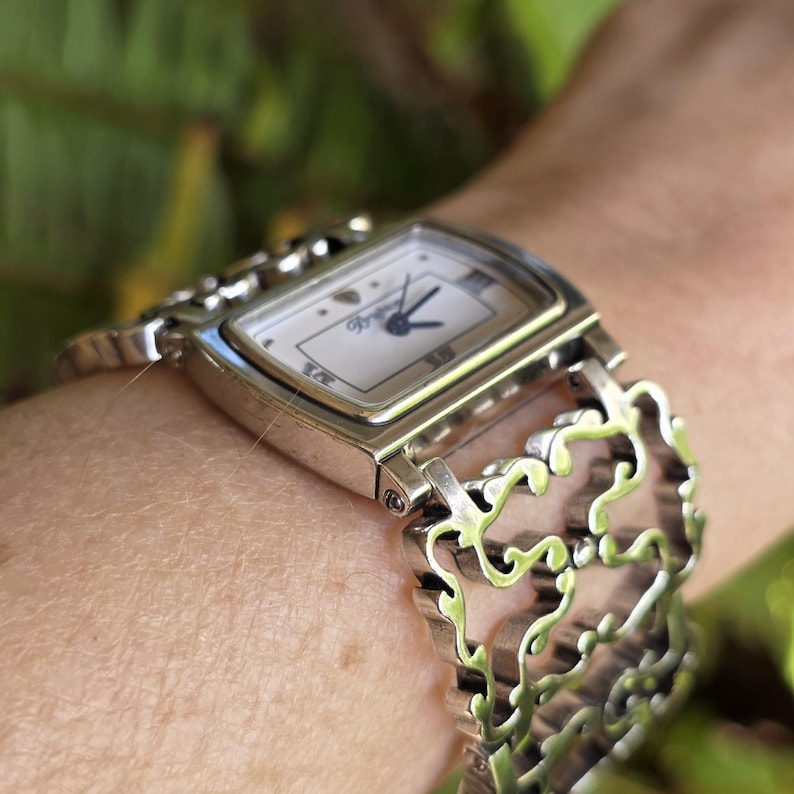May include: A silver-toned wristwatch with a rectangular face and a decorative, open-link band. The watch face is white with black hands and the brand name "Brighton". The watch is worn on a wrist.