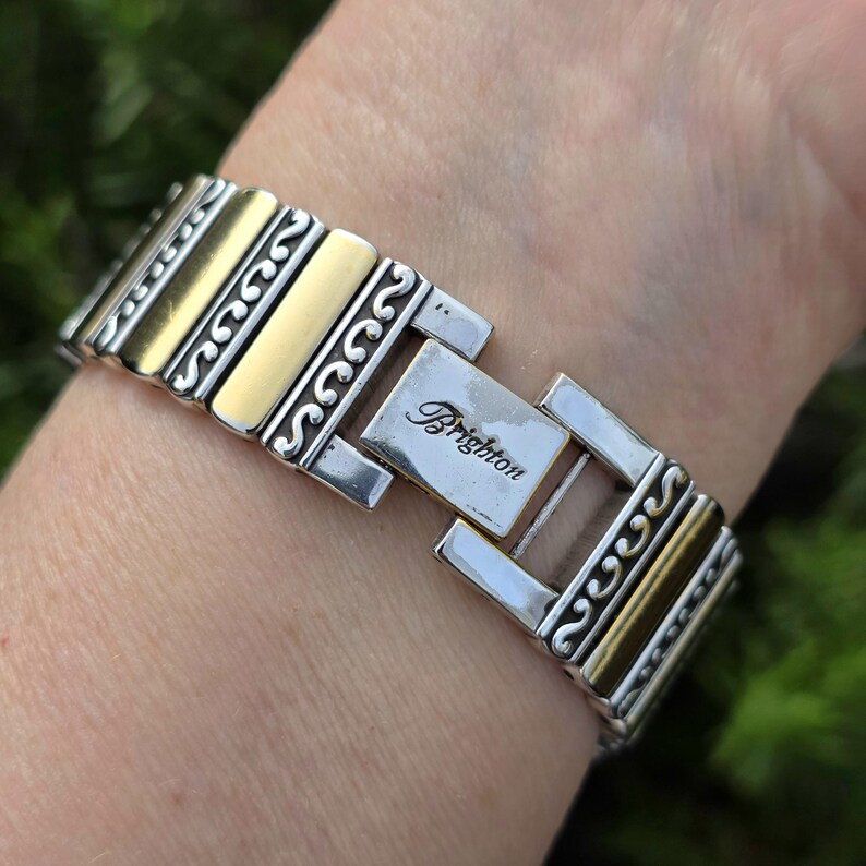 Op de afbeelding: Zilver- en goudkleurige metalen armband met de merknaam "Brighton" gegraveerd op de sluiting. De armband heeft een decoratief ontwerp met afwisselend zilveren en gouden rechthoekige schakels. De armband zit om een pols.