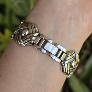 May include: A silver-toned metal link bracelet featuring repeating square-shaped segments with a woven, crisscross pattern and a central polished sphere. The bracelet includes a rectangular clasp mechanism connecting the decorative metal links.