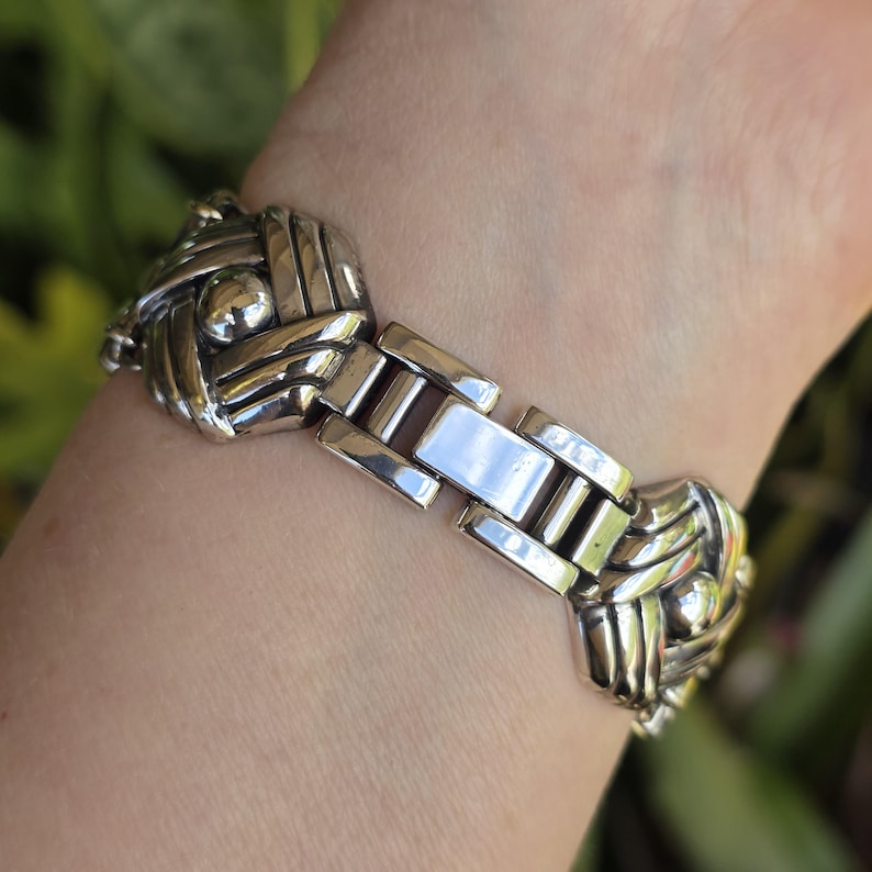 May include: A silver-toned metal link bracelet featuring repeating square-shaped segments with a woven, crisscross pattern and a central polished sphere. The bracelet includes a rectangular clasp mechanism connecting the decorative metal links.
