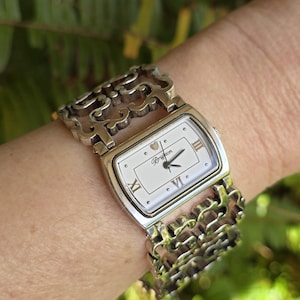 May include: Silver-toned Brighton watch with a rectangular white face and Roman numeral hour markers. The watch features an ornate, open-link bracelet. The watch hands are black, and the watch face has the Brighton logo.