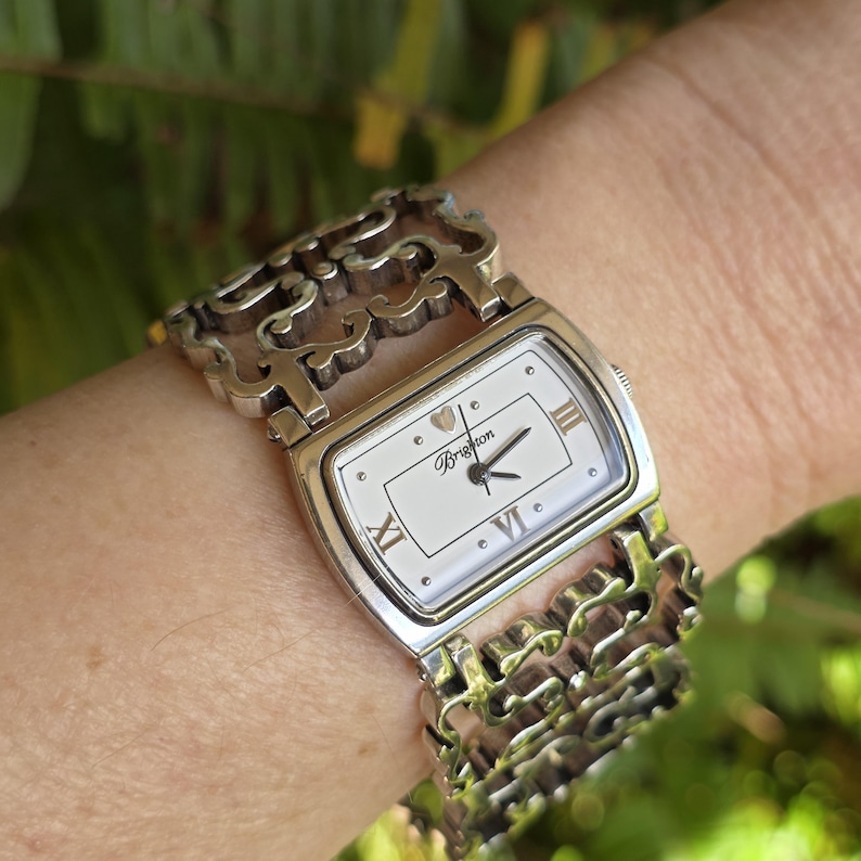 May include: Silver-toned Brighton watch with a rectangular white face and Roman numeral hour markers. The watch features an ornate, open-link bracelet. The watch hands are black, and the watch face has the Brighton logo.