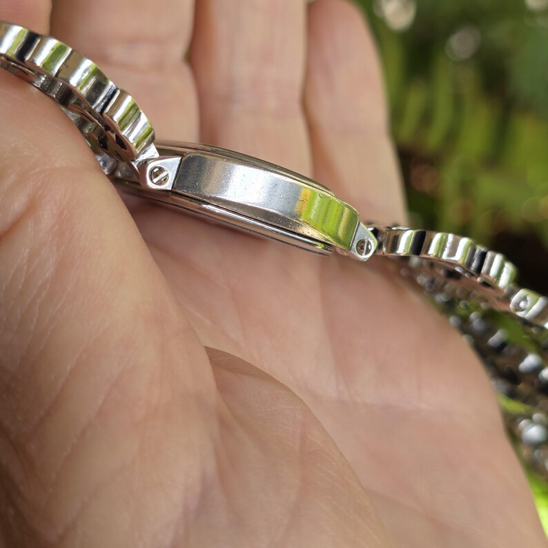 May include: A silver wristwatch with a rectangular face and a matching, patterned band. The watch is held in a hand, with a blurred green background. The watch band has a repeating, scalloped design.