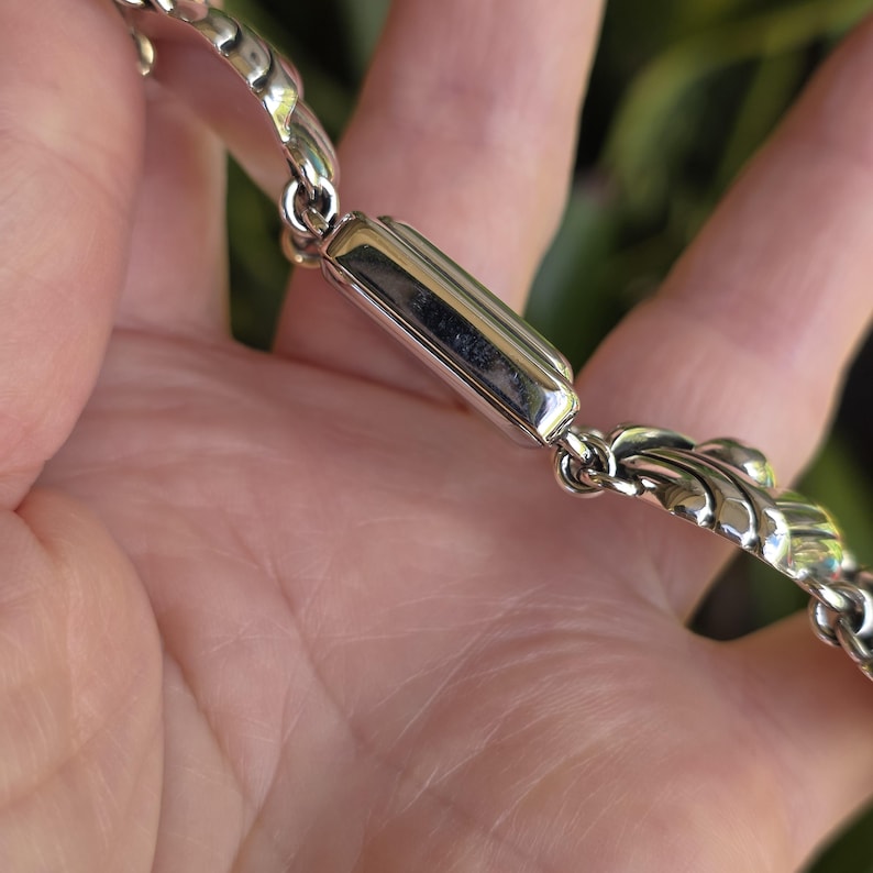 May include: A silver-toned metal bracelet featuring a series of interconnected links. The design includes a smooth, rectangular central bar element joined to textured, scalloped-edge links that create a continuous chain pattern with a polished finish.