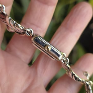 May include: A silver-toned rectangular wristwatch case with a small gold-toned crown on the side. The watch is attached to a decorative silver-toned metal link bracelet featuring a repeating curved, shell-like pattern throughout the band.