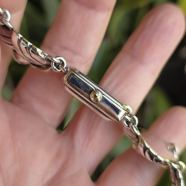 May include: A silver-toned rectangular wristwatch case with a small gold-toned crown on the side. The watch is attached to a decorative silver-toned metal link bracelet featuring a repeating curved, shell-like pattern throughout the band.