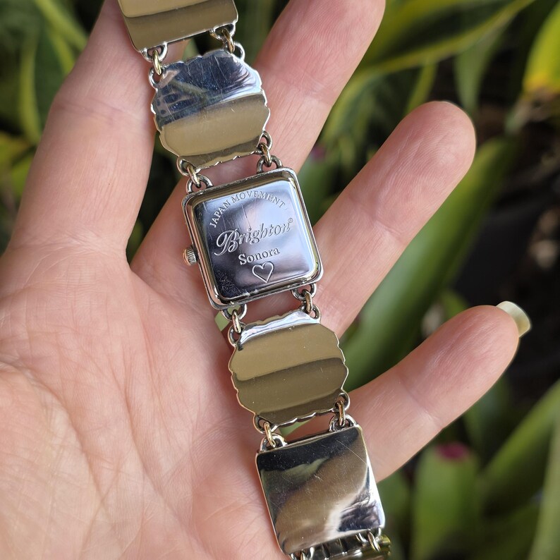 May include: A silver-tone link watch with a square case featuring the text Brighton, Sonora, and a heart symbol engraved on the back. The watch case is marked Japan Movement. The band consists of polished, scalloped-edge metal links connected by rings.