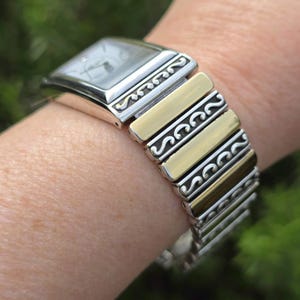 Op de afbeelding: Een zilver- en goudkleurig horloge met een rechthoekige wijzerplaat en een decoratieve band. De wijzerplaat is zilverkleurig met zwarte wijzers. De band wisselt af tussen zilveren en goudkleurige metalen segmenten, met een sierlijk ontwerp.