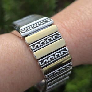 Op de afbeelding: Zilver- en goudkleurig horloge met een rekbare band. De wijzerplaat is rechthoekig. De band heeft afwisselend goudkleurige en zilverkleurige schakels met decoratieve krullen. Het horloge wordt om de pols gedragen.