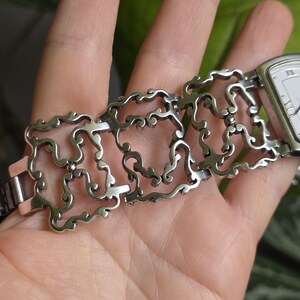 May include: Silver-toned watch with a rectangular face and Roman numeral hour markers. The watch band features an intricate, openwork design with a scrolling, ornamental pattern. The watch is shown on a person's hand.