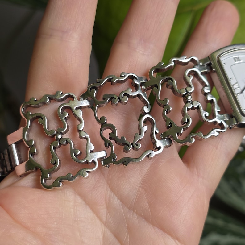May include: Silver-toned watch with a rectangular face and Roman numeral hour markers. The watch band features an intricate, openwork design with a scrolling, ornamental pattern. The watch is shown on a person's hand.