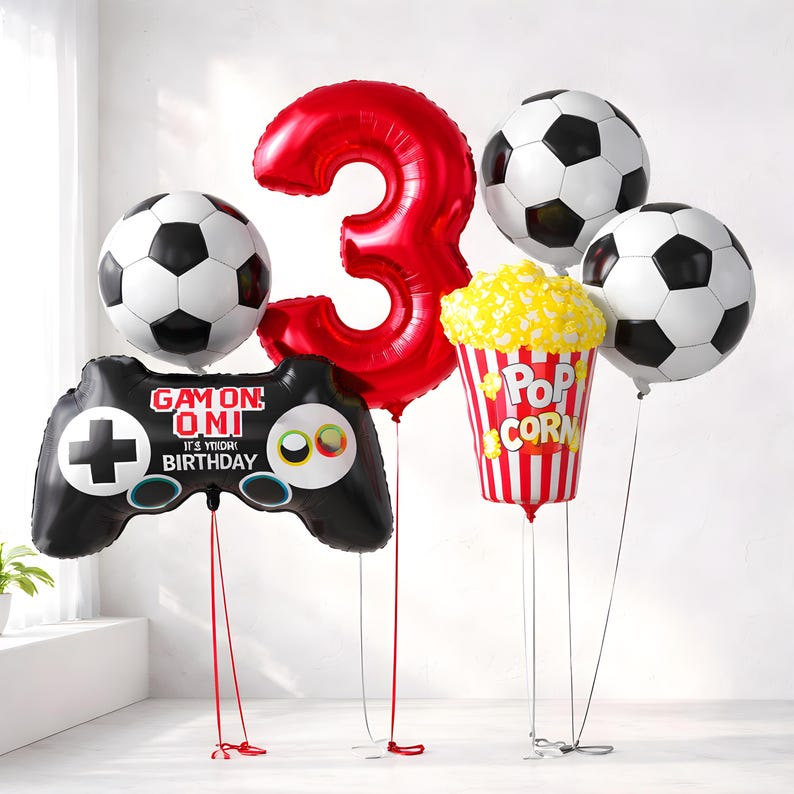 May include: A selection of birthday balloons. Features a large red number 3 balloon, three football balloons, a black video game controller balloon with text, and a popcorn balloon.