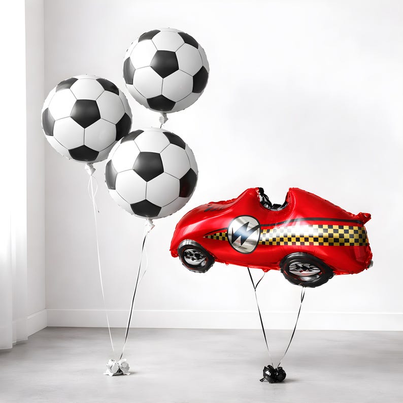 May include: Three football-shaped balloons with black and white panels, and a red race car balloon with black wheels and a chequered flag design. The balloons are tied to weights with ribbons, ideal for a sports-themed party.