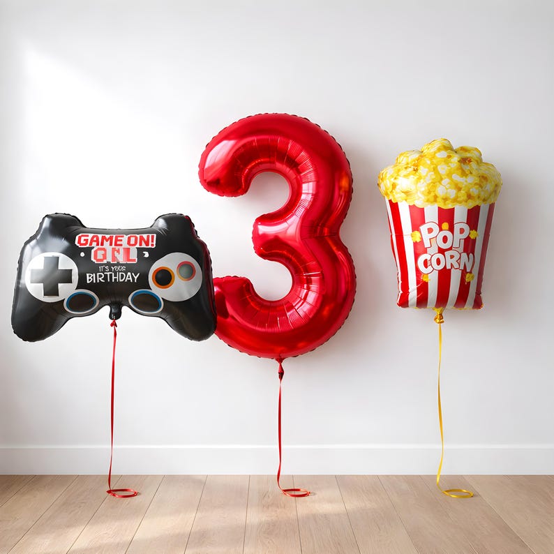 May include: A celebratory image featuring three foil balloons. A large red number 3 balloon is positioned between a black video game controller balloon with the text "GAME ON! IT'S YOUR BIRTHDAY" and a popcorn-shaped balloon.