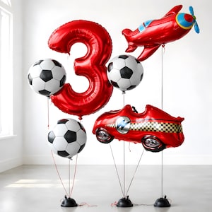 May include: A display of red foil balloons, featuring a large number 3, footballs, a race car, and an aeroplane. The balloons are set on black bases with red strings, ideal for a birthday party.