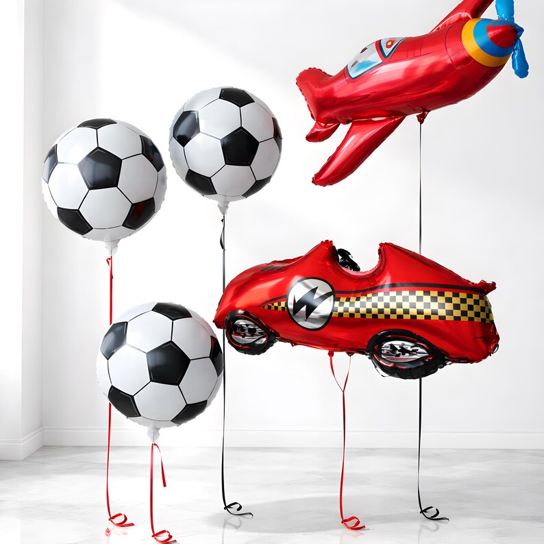 May include: A selection of foil balloons shaped like footballs, a red racing car, and a red aeroplane. The footballs are white with black pentagon shapes. The car and aeroplane are red with black and silver details.