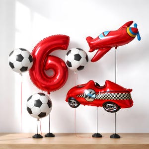 May include: A collection of red foil balloons, including the number 6, footballs, a plane, and a race car. The balloons are displayed on black stands, ideal for a birthday celebration.