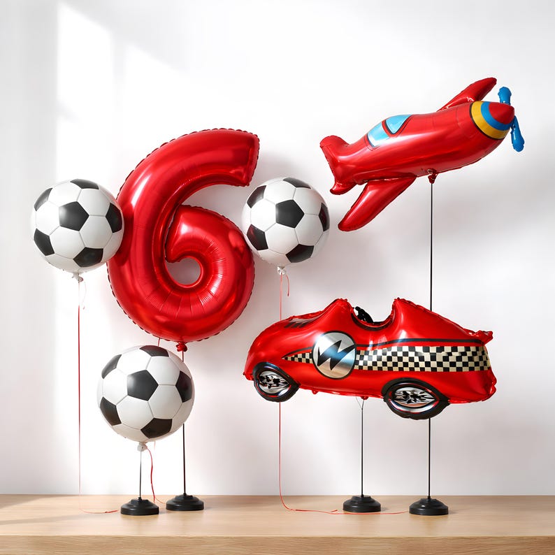 May include: A collection of red foil balloons, including the number 6, footballs, a plane, and a race car. The balloons are displayed on black stands, ideal for a birthday celebration.