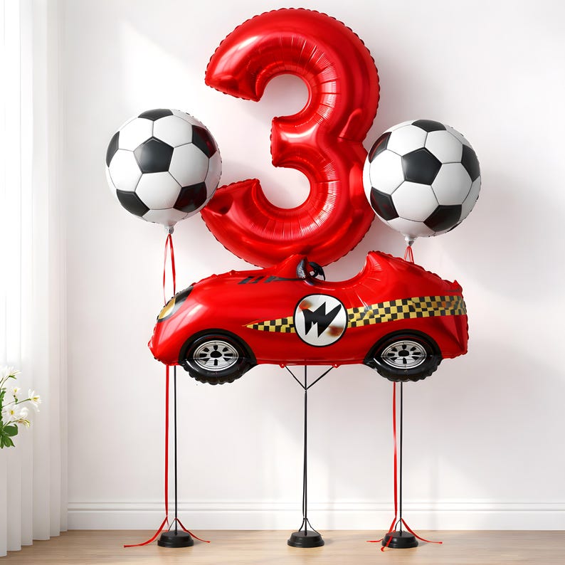 May include: A celebratory balloon display for a third birthday. It includes a large red number 3 balloon, a red race car balloon, and two football-shaped balloons. The race car has a chequered flag design and a white emblem.
