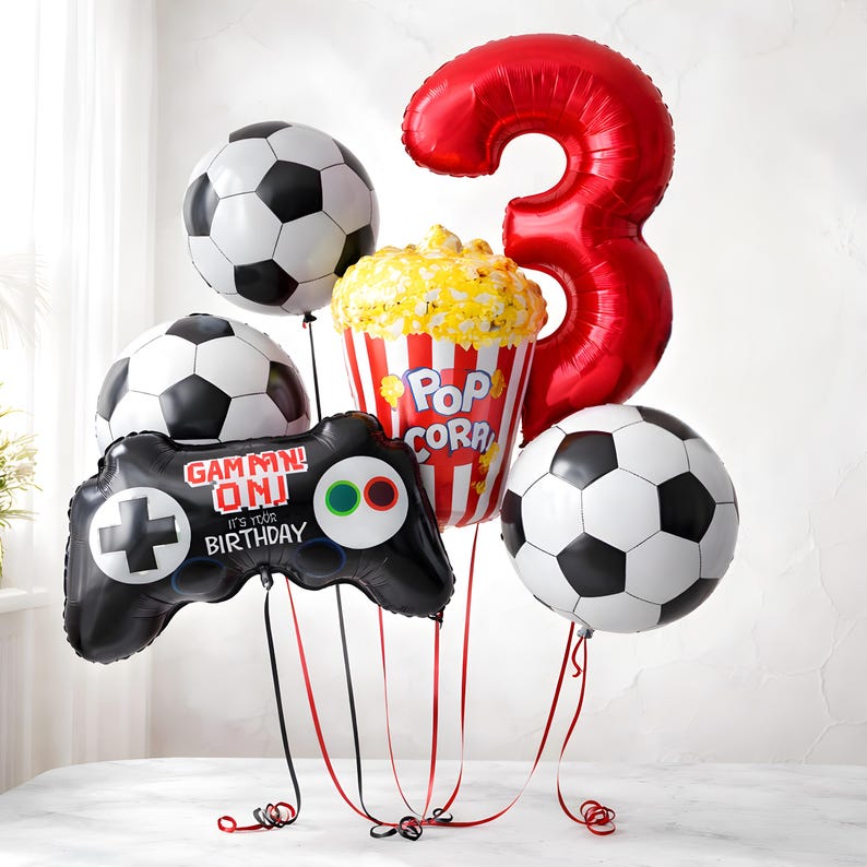 May include: A celebratory balloon arrangement with football, popcorn, and controller-shaped balloons. A large red number 3 balloon is also present. The controller balloon reads "GAMMN! ON! IT'S YOUR BIRTHDAY". Black and red ribbons are attached.