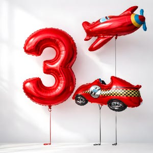 May include: Three red foil balloons: a number 3, a race car, and an aeroplane. The race car and aeroplane have black strings. The aeroplane has a blue, yellow, and red propeller. The race car features a black and yellow chequered pattern.