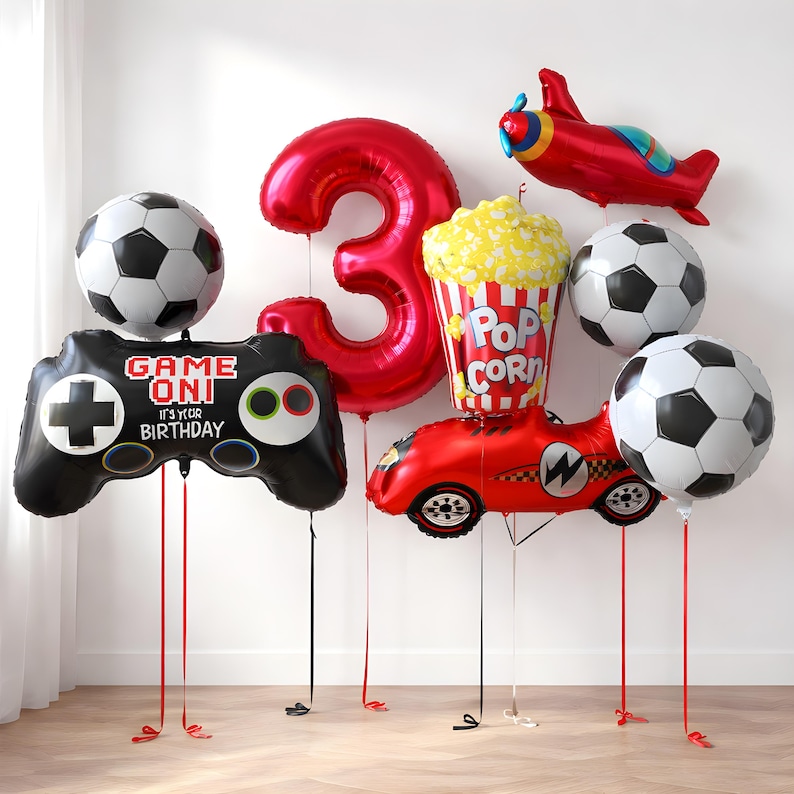 May include: A selection of birthday balloons, including a large red number 3, footballs, a video game controller, a race car, a popcorn container, and a red aeroplane. The video game controller balloon has the text "GAME ON! IT'S YOUR BIRTHDAY".
