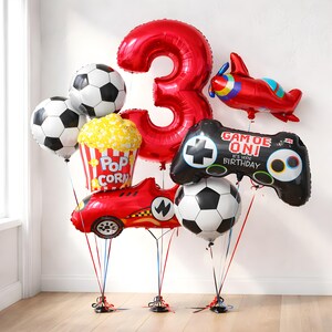 May include: A selection of birthday balloons, including a large red number 3, footballs, a popcorn container, a race car, a video game controller, and a red aeroplane. The controller reads "Game On! It's Your Birthday."