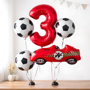 May include: A celebratory balloon display with a large red number 3, three football-shaped balloons, and a red racing car balloon. The balloons are black and white, and the car has a chequered flag design. Ideal for a birthday party.