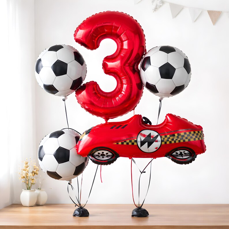 May include: A celebratory balloon display with a large red number 3, three football-shaped balloons, and a red racing car balloon. The balloons are black and white, and the car has a chequered flag design. Ideal for a birthday party.