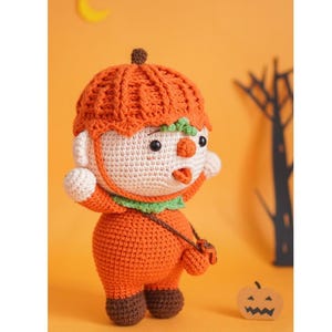 May include: A crocheted pumpkin-themed plush toy wearing an orange hat and holding a small orange bag. The toy has a white face with black eyes and a red nose. The toy is standing on a yellow surface with a small black and orange pumpkin in the background.