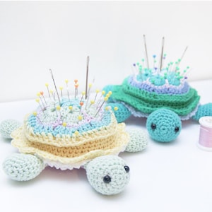 Op de afbeelding: Twee handgemaakte gehaakte schildpadspeldenkussens. Eén is lichtblauw, geel en wit, de andere is groen, blauw en paars. Beide hebben meerdere spelden ingebracht. De schildpadden hebben kleine, ronde hoofden en vier vinachtige voeten.