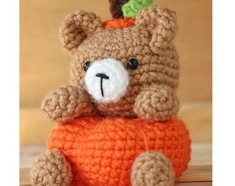 May include: A crocheted brown bear wearing an orange pumpkin costume. The bear has a white nose and black eyes. The pumpkin has a green stem.