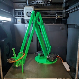 May include: A green 3D printed robotic arm with a circular base and two servo motors. The arm is inside a 3D printer with a white build plate that reads "Build Volume: 256 x 256 x 256 mm^3".