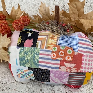 May include: A handmade patchwork pumpkin with a wooden stem, crafted from various fabric scraps in a variety of colors and patterns. The pumpkin is decorated with dried foliage and orange decorative berries, perfect for fall decor.
